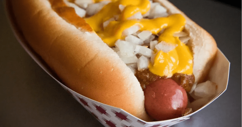 A hot dog in a bun topped with mustard, cheese, and diced onions, served in a paper tray.