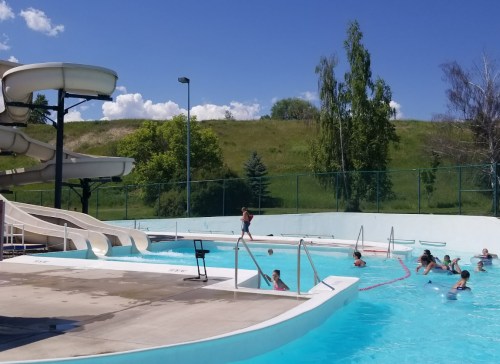 A sunny outdoor pool with water slides, children swimming, and green hills in the background.