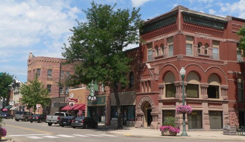 Historic buildings line a street, featuring red brick architecture and storefronts, with trees and flowers adding color.