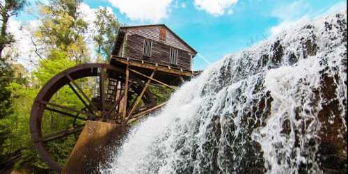 A historic watermill with a large wooden wheel, surrounded by lush greenery and cascading water under a blue sky.