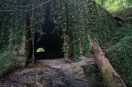 A stone tunnel covered in ivy, with a small stream flowing in front and a circular opening at the far end.