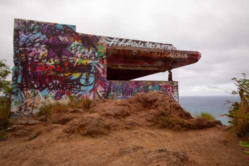 A graffiti-covered concrete structure on a rocky hillside overlooking the ocean under a cloudy sky.