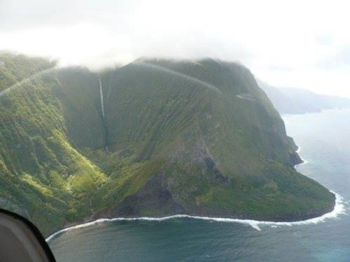 Aerial view of lush green cliffs meeting the ocean, with a waterfall cascading down the mountainside.
