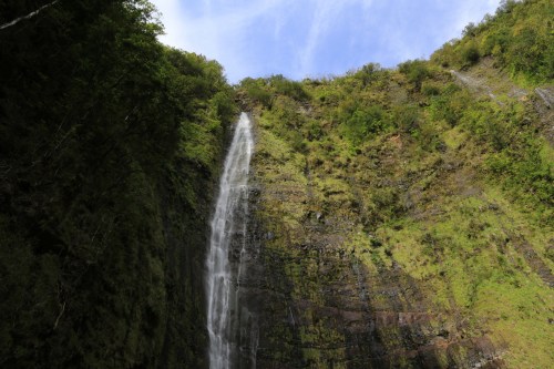 A tall waterfall cascading down a rocky cliff, surrounded by lush greenery and a clear blue sky.