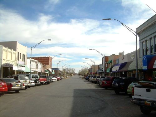 A quiet street lined with parked cars and storefronts under a blue sky with scattered clouds.