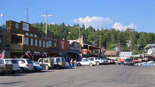 A small town street lined with wooden buildings and parked cars, surrounded by trees under a clear blue sky.