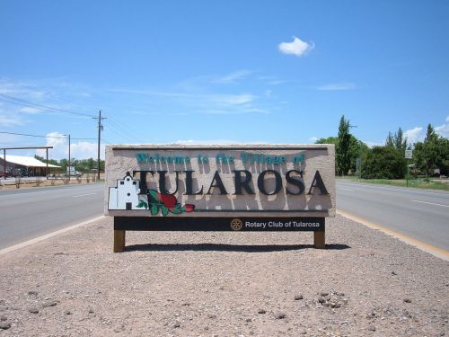 Welcome sign for the Village of Tularosa, featuring a rose and a small building illustration. Clear blue sky above.