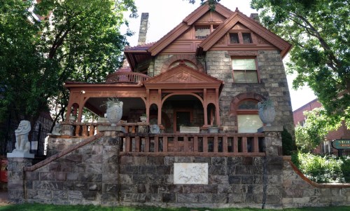 A historic stone house with a red wooden porch, surrounded by trees and decorative sculptures in the yard.