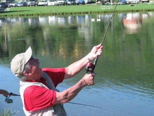 An older man in a red shirt and cap is fishing by a calm lake, casting his line with enthusiasm.