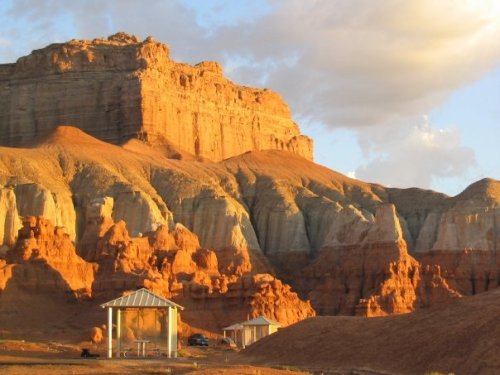 Sunlit rock formations with layered colors and a small gazebo in the foreground against a dramatic sky.