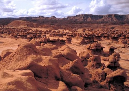A vast desert landscape featuring unique rock formations and distant mesas under a cloudy sky.