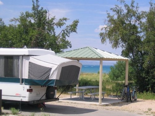 A camper parked near a picnic area with a gazebo and bicycles, overlooking a scenic lake and greenery.