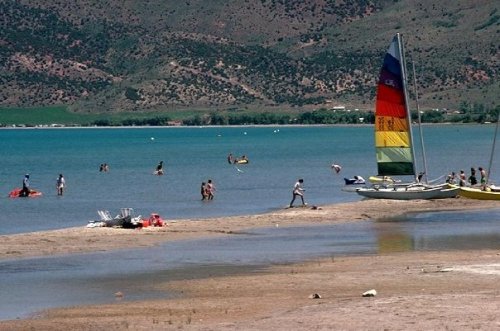 A sunny beach scene with people swimming, sailing, and enjoying the water, featuring a colorful sailboat in the foreground.