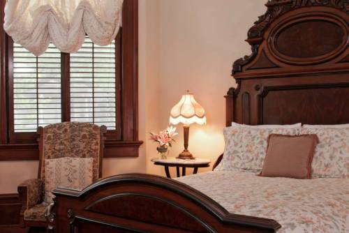 Cozy bedroom featuring a wooden bed, floral bedding, a vintage armchair, and a lamp beside a window with blinds.