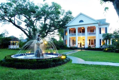 A grand white mansion with columns, surrounded by lush greenery and a fountain in the foreground, at dusk.