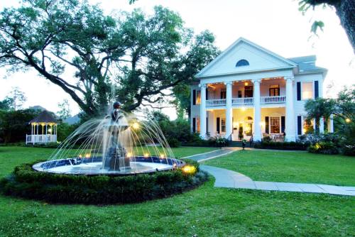 A grand white mansion with columns, surrounded by lush greenery and a fountain in the foreground, at dusk.