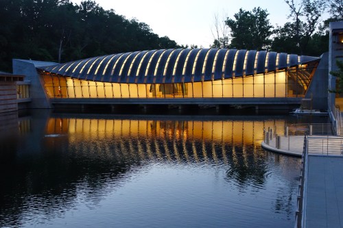 A modern building with a curved roof reflects in a calm lake at dusk, surrounded by trees.
