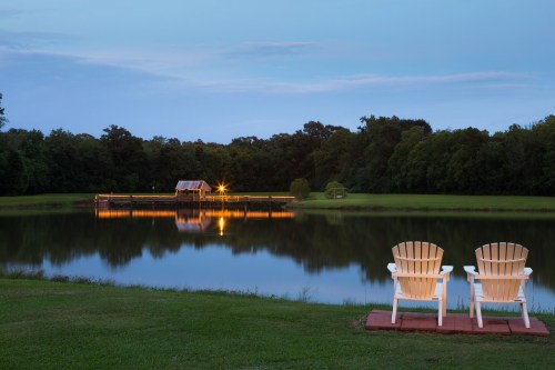 Two white Adirondack chairs sit by a calm lake at dusk, reflecting a cozy cabin and surrounding trees.