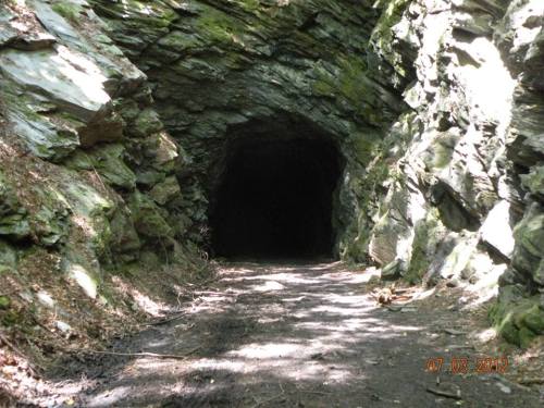 A dark tunnel entrance surrounded by rocky walls and scattered sunlight on a dirt path.