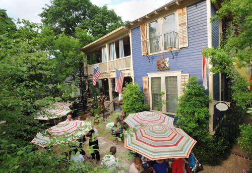 A charming blue house surrounded by greenery, featuring outdoor seating with colorful umbrellas and American flags.