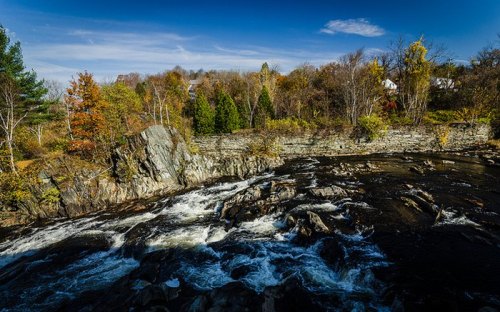 A scenic river flows over rocks, surrounded by autumn trees and a clear blue sky.