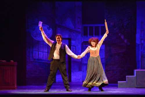 Two performers on stage, holding hands and celebrating, with a colorful backdrop of a theatrical set.