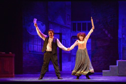 Two performers on stage, holding hands and celebrating, with a colorful backdrop of a theatrical set.