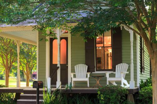 A charming green cottage with a porch, white chairs, and large windows surrounded by trees and greenery.