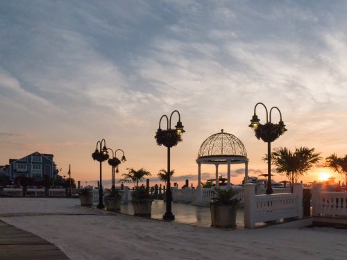 A beachside gazebo with decorative lights at sunset, surrounded by palm trees and a charming building in the background.