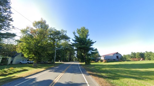 A quiet rural road lined with trees, leading to a grassy area and a barn under a clear blue sky.