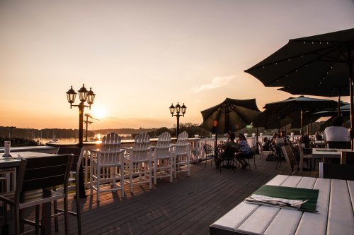 A scenic outdoor dining area with tables and chairs, overlooking a sunset by the water.