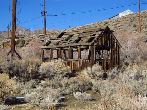 A dilapidated wooden structure stands in a barren landscape, surrounded by dry shrubs and power lines under a clear blue sky.