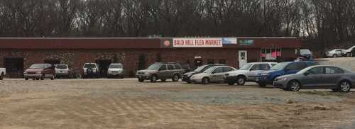 Exterior view of Bald Hill Flea Market with parked cars and a gravel lot, surrounded by trees in the background.