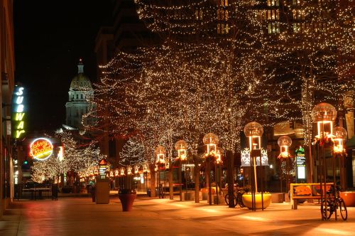 A festive street at night, lined with trees adorned in twinkling lights and illuminated storefronts.