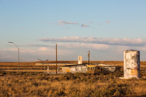Abandoned building and water tower in a desolate landscape under a clear sky, with sparse vegetation and distant hills.