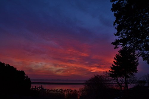 Vibrant sunset over a calm lake, with colorful clouds and silhouettes of trees along the shoreline.