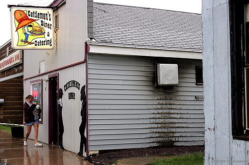A woman walks past Cattlemen's Diner, featuring a colorful sign and a gray exterior, on a rainy day.