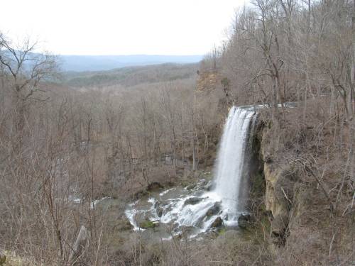 A waterfall cascades down rocky cliffs, surrounded by bare trees and a distant mountain landscape.