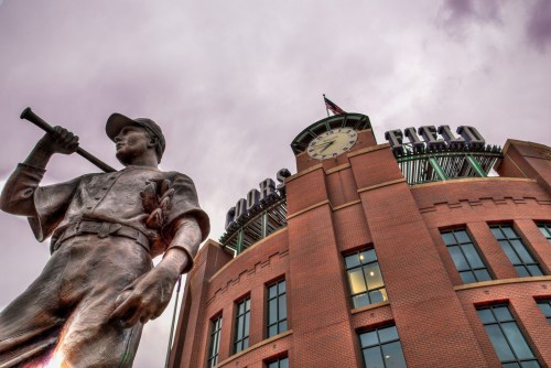 A bronze statue of a baseball player stands in front of Coors Field, with a cloudy sky above.