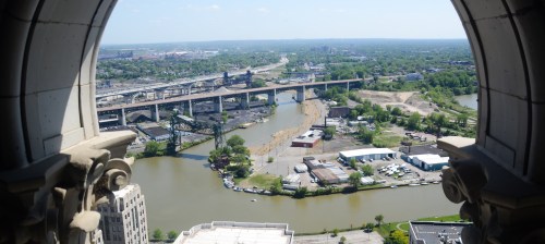 Aerial view of a river and industrial area, framed by an ornate architectural window. Green landscape and bridges visible.
