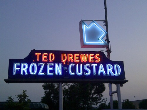 Neon sign for Ted Drewes Frozen Custard, featuring bright blue and red lettering against a twilight sky.