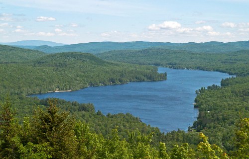 A scenic view of a winding lake surrounded by lush green forests and distant mountains under a blue sky.