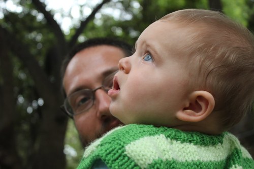 A close-up of a baby with blue eyes looking up, wearing a green sweater, with a blurred adult figure in the background.