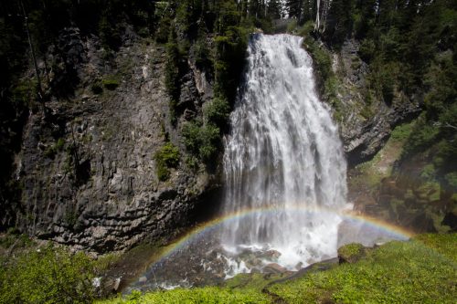 A stunning waterfall cascades down rocky cliffs, creating a vibrant rainbow over the misty pool below.