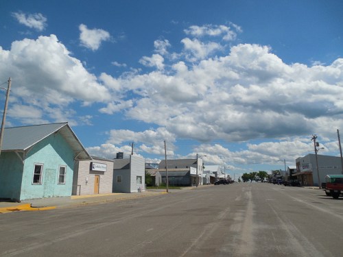 A quiet street in a small town, lined with buildings under a blue sky filled with fluffy white clouds.