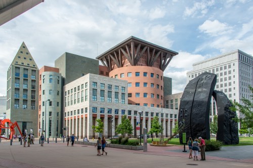 Modern architectural buildings with unique designs, surrounded by greenery and people walking in an urban setting.