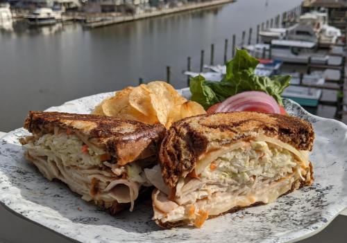 A close-up of a turkey sandwich with coleslaw, served with chips and lettuce, overlooking a marina.