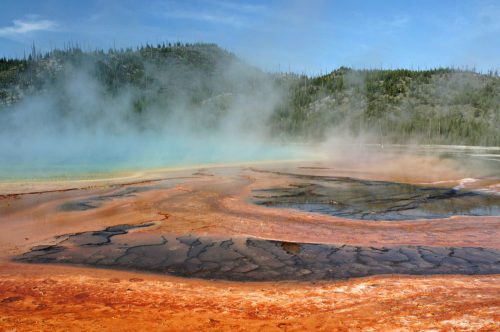 A colorful geothermal area with steam rising, featuring vibrant orange and blue hues against a forested backdrop.