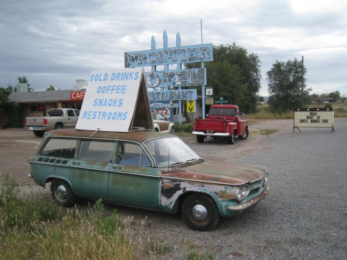 A vintage car with a sign for cold drinks and snacks parked outside a rustic motel and restaurant.