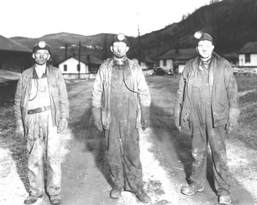 Three coal miners stand on a dirt road, wearing helmets with lights and covered in dirt, with houses and hills in the background.